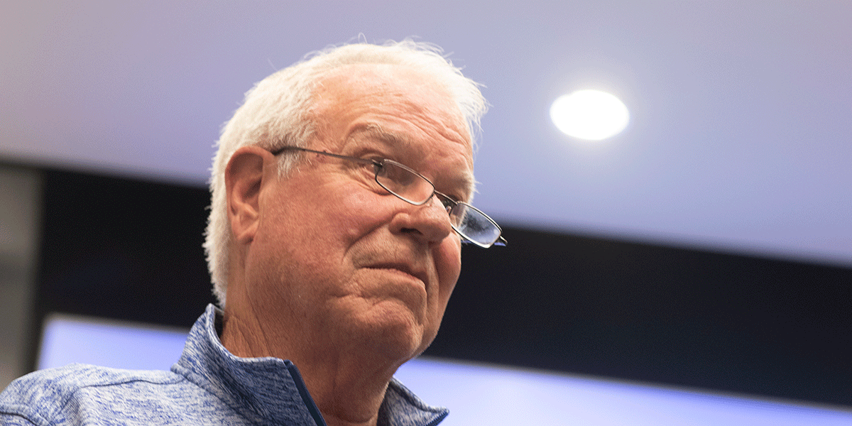 A man with white hair, glasses, and a slightly intense expression is pictured from a medium-close angle beneath his face. A ceiling list is illuminated and visible above him and to the left.